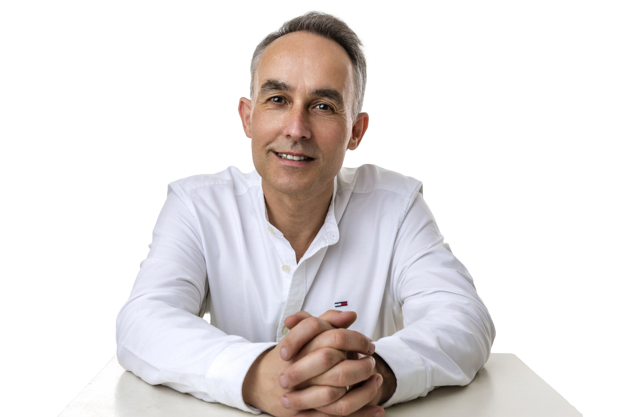 Chris Cooper, wearing a white shirt, hands clasped together on a table, with a welcoming smile.